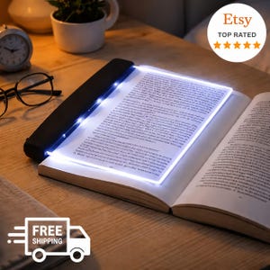 Puede incluir: Un panel de luz de lectura LED rectangular, plano y transparente con un mango de plástico negro en un lado. El dispositivo emite un brillo blanco brillante, iluminando las páginas abiertas de un libro colocado debajo sobre una superficie de madera.
