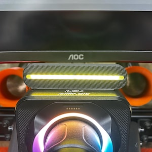 Puede incluir: Primer plano de una configuración de simulador de carreras con una barra de luz LED negra y de fibra de carbono, montada en un dispositivo negro con el texto "SIMAGIC". La configuración incluye componentes naranjas y un monitor curvo.