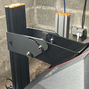 Puede incluir: Un marco de impresora 3D negro y naranja con una superficie gris texturizada. La impresora está montada sobre un soporte de metal negro con brazos ajustables. El soporte está fijado a una pared de bloques de hormigón.