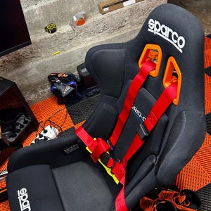 Puede incluir: Un asiento de carreras Sparco negro con diseño de respaldo alto y pasadores de arnés naranjas integrados. El asiento incluye un arnés de carreras rojo brillante de varios puntos, la marca Sparco en blanco y un acabado de tela texturizada.
