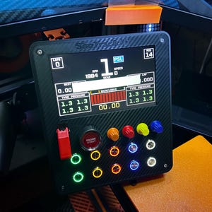 May include: A black carbon fiber sim racing dashboard display with a digital screen showing telemetry data, including RPM, gear, and tire pressure. The unit features a red toggle switch, an engine start button, and several colorful rotary knobs.