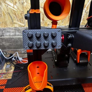 Puede incluir: Un panel de control negro con textura de fibra de carbono y ocho botones circulares pequeños, montado en un marco de metal negro. Encima hay un componente naranja en forma de embudo y debajo un soporte naranja con el texto imTrack.