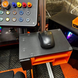 May include: A black Hama computer mouse rests on a carbon fiber textured platform attached to an orange 3D-printed mount. Above, a racing simulator dashboard features an All In One Dash 2.0 screen, an orange Engine Start button, and colorful knobs.