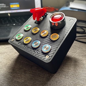 May include: A black button box controller with a carbon fiber texture finish, featuring a red flip-up safety switch, a large red Engine Start button, and seven smaller illuminated push buttons. The front panel displays the text SimTrack3D.