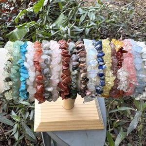 May include: A selection of gemstone chip bracelets in a variety of colours, including green, blue, red, and white. The bracelets are strung together and displayed on a cylindrical, off-white stand. The background features green foliage.