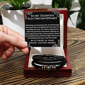 May include: A black leather bracelet with silver and gold accents, presented in a red wooden gift box. The box contains a card with the text "TO MY TALENTED ELECTRICIAN [NAME]" and a heartfelt message.