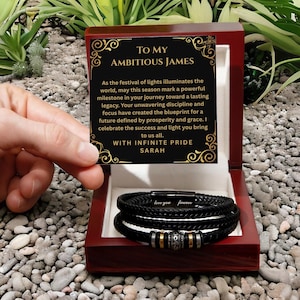 May include: A black braided leather bracelet with a magnetic clasp engraved with "love you forever" and metallic accents, presented in a dark wood box with a card reading "To My Ambitious James" and a message about prosperity and legacy from Sarah.