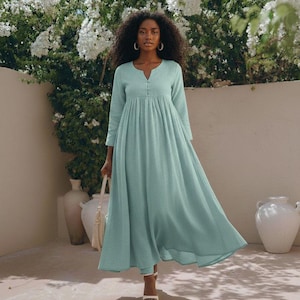 Puede incluir: Un vestido largo y fluido de color azul claro, con mangas largas y escote en V. El vestido tiene la cintura fruncida y llega hasta los tobillos. La modelo lleva pendientes de aro dorados y sandalias blancas. Un bolso blanco se sostiene en la mano izquierda.