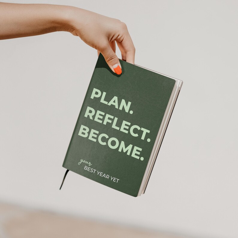 May include: A dark green planner with a hard cover featuring the light green text PLAN. REFLECT. BECOME. and the smaller white text your BEST YEAR YET. The planner has a black ribbon bookmark extending from the bottom edge of the spine.