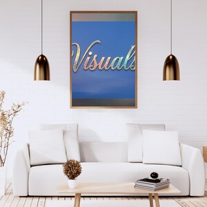 May include: A framed artwork with the word "Visuals" in a colorful, glittery script against a blue background hangs above a white sofa. The room features a light wood coffee table, a white rug, and gold pendant lights.