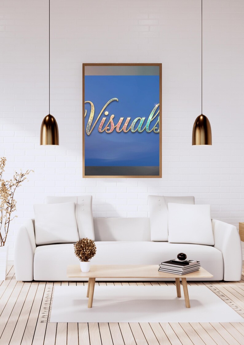 May include: A framed artwork with the word "Visuals" in a colorful, glittery script against a blue background hangs above a white sofa. The room features a light wood coffee table, a white rug, and gold pendant lights.