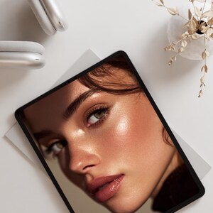 May include: A tablet displaying a close-up of a woman's face with radiant skin and soft makeup. The image is set on a white surface, with a pair of white headphones and a small vase with dried plants in the background.