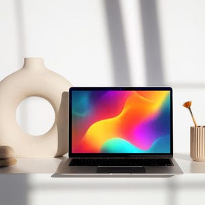 May include: A silver laptop with a vibrant, colorful abstract design on the screen sits on a white surface. Beside it is a beige vase with a circular opening and a small, cylindrical container holding a paintbrush.