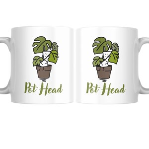 May include: White ceramic mug featuring a green and brown illustration of a plant in a pot, with the text "Pot Head" in green. The mug has a curved handle and is suitable for hot or cold drinks.