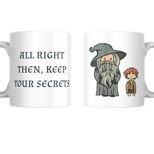 May include: White ceramic mug. One side has the text "ALL RIGHT THEN, KEEP YOUR SECRETS". The other side features a cartoon illustration of a wizard and a hobbit. The mug has a curved handle.