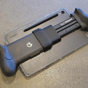 以下が含まれることがあります： 透明な保護ケースに入ったグレーのSamsungタブレット。背面には黒いスタイラスペンが装着されています。テクスチャ加工されたグリップと円形のロゴが付いた黒いプラスチック製のゲームコントローラーマウントが、タブレットの中央に水平に取り付けられています。