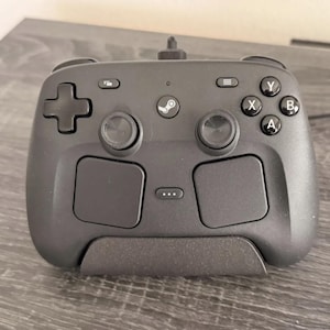 May include: A matte black video game controller featuring a central Steam logo, two analogue sticks, a directional pad, four action buttons labelled A, B, X, and Y, two large touchpads, and a small button with three dots located between the touchpads.