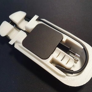 May include: A white 3D-printed plastic cable organiser tray with a rectangular cutout holding a black square power adapter and a black charging cable. The organiser features moulded slots to secure the cable and the adapter in a compact arrangement.