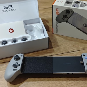 Puede incluir: Un mando de juego móvil gris con joysticks negros, una cruceta y botones de acción azules. El dispositivo se muestra con su caja de venta blanca etiquetada como G8 Galileo y un pequeño inserto blanco que contiene tres tapas de joystick.