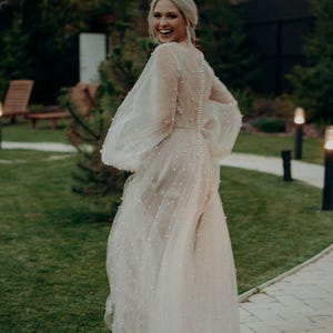 May include: A pale wedding dress with sheer, puffed sleeves and a long, flowing skirt, decorated with small embellishments. The bride smiles as she walks on a stone path in a garden, surrounded by greenery and soft lighting, creating a romantic atmosphere.