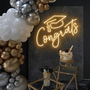May include: A glowing yellow neon sign displaying the word Congrats with a graduation cap icon above it. A large balloon arch in white, silver, and gold surrounds the sign. A small cake and graduation-themed table decor are present.