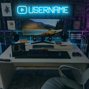 Pode incluir: Uma configuração de jogos moderna com um monitor grande, teclado, rato e laptop numa secretária branca. Uma placa de néon com a palavra "USERNAME" está acima do monitor. A sala tem uma parede escura e texturizada e vários acessórios tecnológicos.
