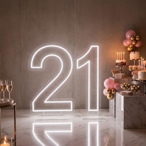 May include: A large, illuminated neon sign displaying the number 21 in bright white light, standing on clear acrylic bases. The display is surrounded by pink and gold balloons, tiered cakes with lit candles, and champagne glasses on a marble floor.