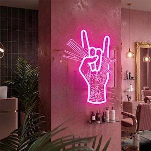 Puede incluir: Un letrero de neón rosa que representa una mano haciendo el gesto de rock and roll, sosteniendo tijeras y un peine, con tatuajes florales. El letrero está montado en una pared rosa, lo que sugiere un tema de salón o barbería.