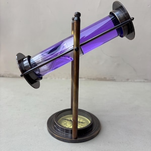 May include: A vintage brass and purple glass instrument with a compass base. The glass tube is filled with a vibrant purple liquid and is held in place by a dark metal frame. The compass is set in a circular brass base.