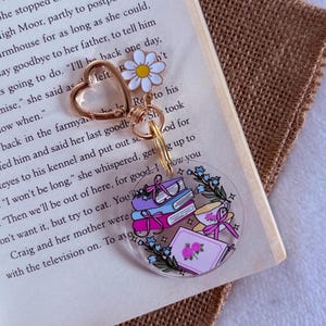 May include: A clear acrylic keyring with a design of books, a teacup, and flowers. The keyring has a gold-coloured heart-shaped clasp and a daisy charm. The keyring is resting on an open book.