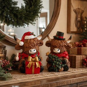 Pode incluir: Duas figuras de vacas Highland castanhas com pelo felpudo. A vaca da esquerda usa um gorro de Pai Natal vermelho e um cachecol vermelho e verde, segurando um presente vermelho com um laço dourado. A da direita usa uma cartola preta e um cachecol.