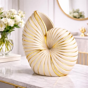 Può includere: Vaso decorativo color crema a forma di conchiglia Nautilus, con dettagli dorati e strass incastonati. Il vaso ha una texture a coste e un'ampia apertura, adatto per composizioni floreali o come elemento decorativo.