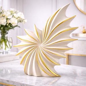 May include: A cream-colored, abstract sculpture with gold accents. The decorative piece has a spiral design with pointed edges and gold lines. The sculpture is placed on a marble surface, with a vase of white flowers in the background.