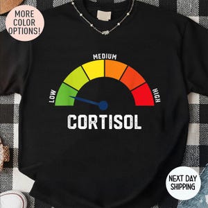 May include: Black sweatshirt with a graphic of a cortisol meter. The meter displays a range from low to high, with a needle pointing towards the low end. The word "CORTISOL" is printed in white below the meter. The shirt also has a "MORE COLOR OPTIONS!" graphic.