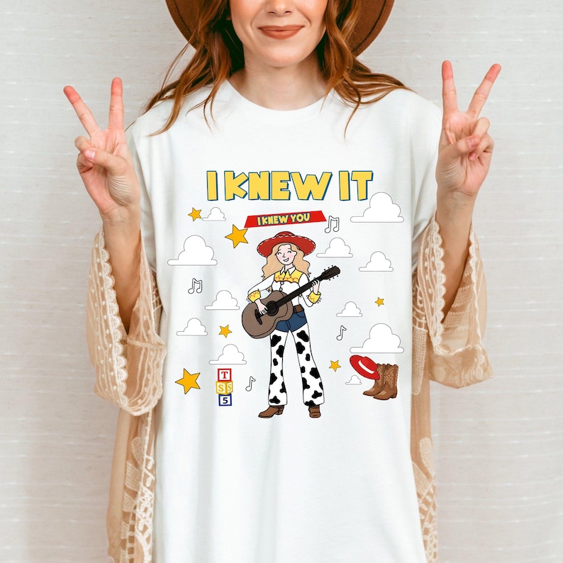 May include: A white t-shirt featuring a graphic of a cowgirl with blonde hair, a red hat, and cow-print pants playing an acoustic guitar. The design includes the text I KNEW IT and I KNEW YOU, surrounded by yellow stars, white clouds, and musical notes.