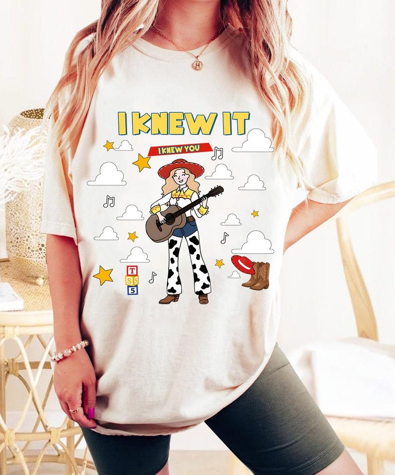 May include: A cream-colored t-shirt featuring a graphic of a blonde cowgirl playing an acoustic guitar. The design includes the text I KNEW IT and I KNEW YOU, surrounded by clouds, stars, musical notes, a red hat, brown boots, and toy blocks.