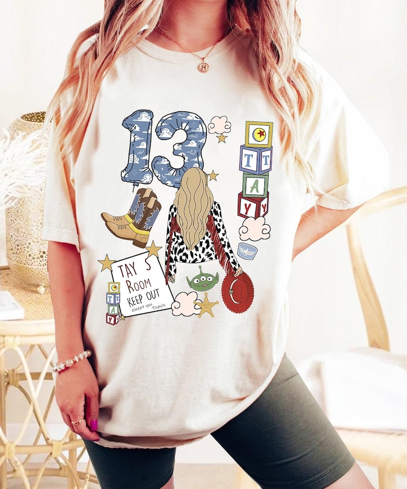 May include: A cream-coloured t-shirt featuring a graphic print of a person with long blonde hair, a cowboy boot, a red hat, a green alien, and stacked blocks spelling TAY. The text reads TAY'S ROOM KEEP OUT except you Travis.