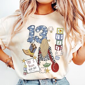 May include: A cream-coloured t-shirt featuring a graphic print of the number 13 in a blue cloud pattern, a blonde person, cowboy boots, alphabet blocks, a green alien, a red hat, and a sign that reads TAY'S ROOM KEEP OUT except you Travis.