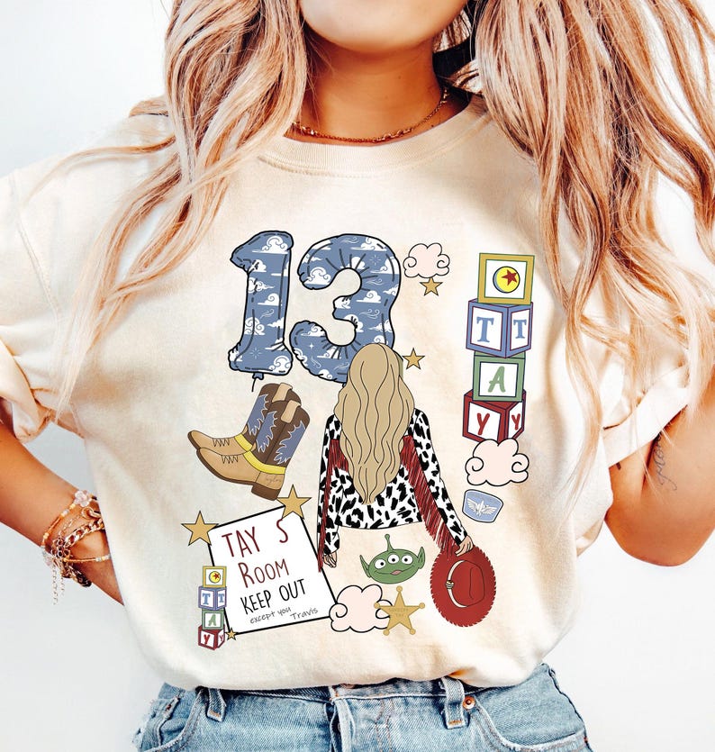 May include: A cream-coloured t-shirt featuring a graphic print of the number 13 in a blue cloud pattern, a blonde person, cowboy boots, alphabet blocks, a green alien, a red hat, and a sign that reads TAY'S ROOM KEEP OUT except you Travis.