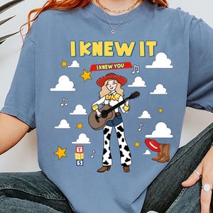 May include: A blue t-shirt featuring a graphic of a cowgirl playing an acoustic guitar, surrounded by white clouds, yellow stars, and musical notes. The text reads I KNEW IT and I KNEW YOU, with a red hat, brown boots, and a block with letters T, S, 5.