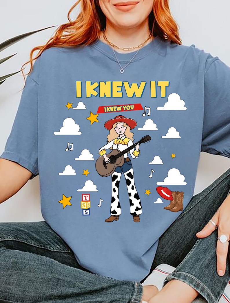 May include: A blue t-shirt featuring a graphic of a cowgirl playing an acoustic guitar, surrounded by white clouds, yellow stars, and musical notes. The text reads I KNEW IT and I KNEW YOU, with a red hat, brown boots, and a block with letters T, S, 5.
