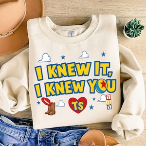 May include: A cream-colored crewneck sweatshirt featuring the blue and yellow text I KNEW IT, I KNEW YOU with a red cowboy hat, brown boots, a red heart with the letters TS, a colorful ball, and a stack of blocks labeled T, S, 5 with blue clouds.
