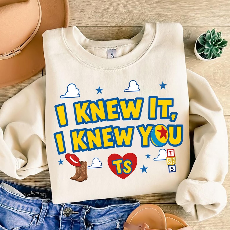 May include: A cream-colored crewneck sweatshirt featuring the blue and yellow text I KNEW IT, I KNEW YOU with a red cowboy hat, brown boots, a red heart with the letters TS, a colorful ball, and a stack of blocks labeled T, S, 5 with blue clouds.