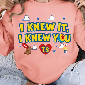 May include: A peach-colored crewneck sweatshirt featuring the text "I KNEW IT, I KNEW YOU" in blue and yellow. The design includes a red heart with "TS" inside, a pair of brown cowboy boots with a red hat, blue stars, white clouds, and toy blocks.
