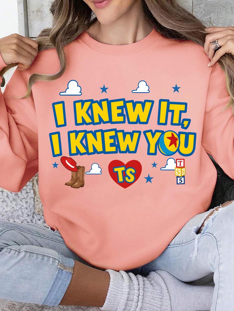 May include: A peach-colored crewneck sweatshirt featuring the text "I KNEW IT, I KNEW YOU" in blue and yellow. The design includes a red heart with "TS" inside, a pair of brown cowboy boots with a red hat, blue stars, white clouds, and toy blocks.