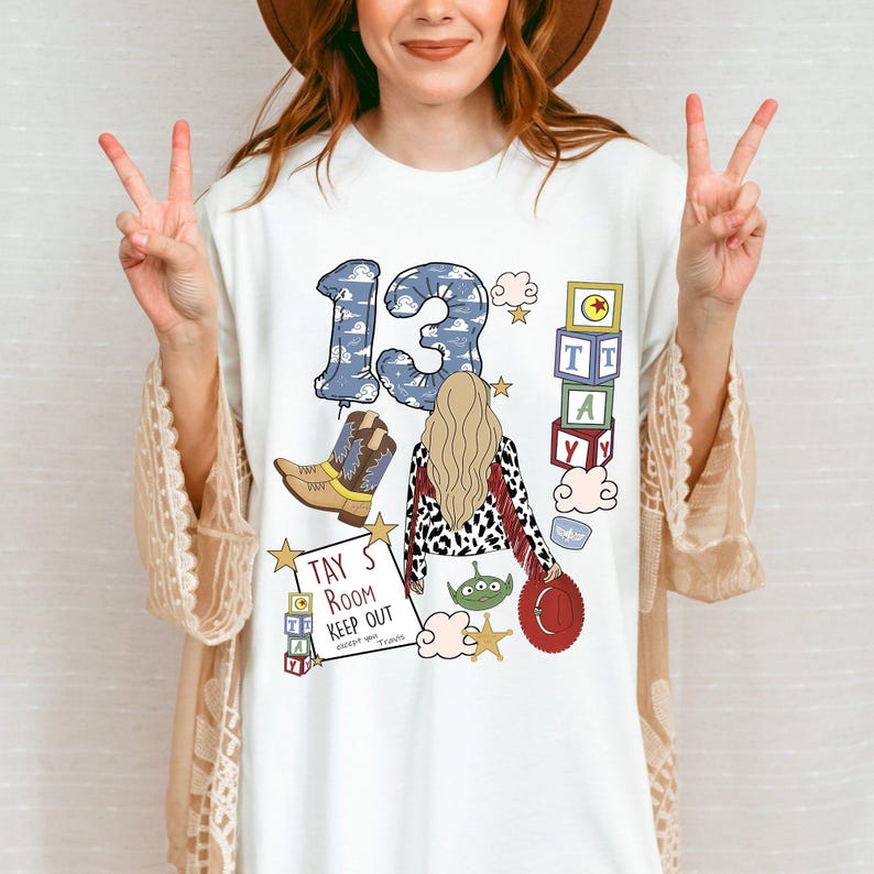May include: A white t-shirt featuring a graphic print with the number 13 in a blue cloud pattern, a blonde character, cowboy boots, alphabet blocks spelling TAY, a sign reading TAY'S ROOM KEEP OUT except you Travis, a green alien, and a red hat.
