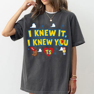 May include: A charcoal gray short-sleeve t-shirt featuring the yellow text I KNEW IT, I KNEW YOU with a ball icon, a red cowboy hat, brown boots, a red heart with TS, blue stars, white clouds, and a stack of alphabet blocks labeled T, S, 5.