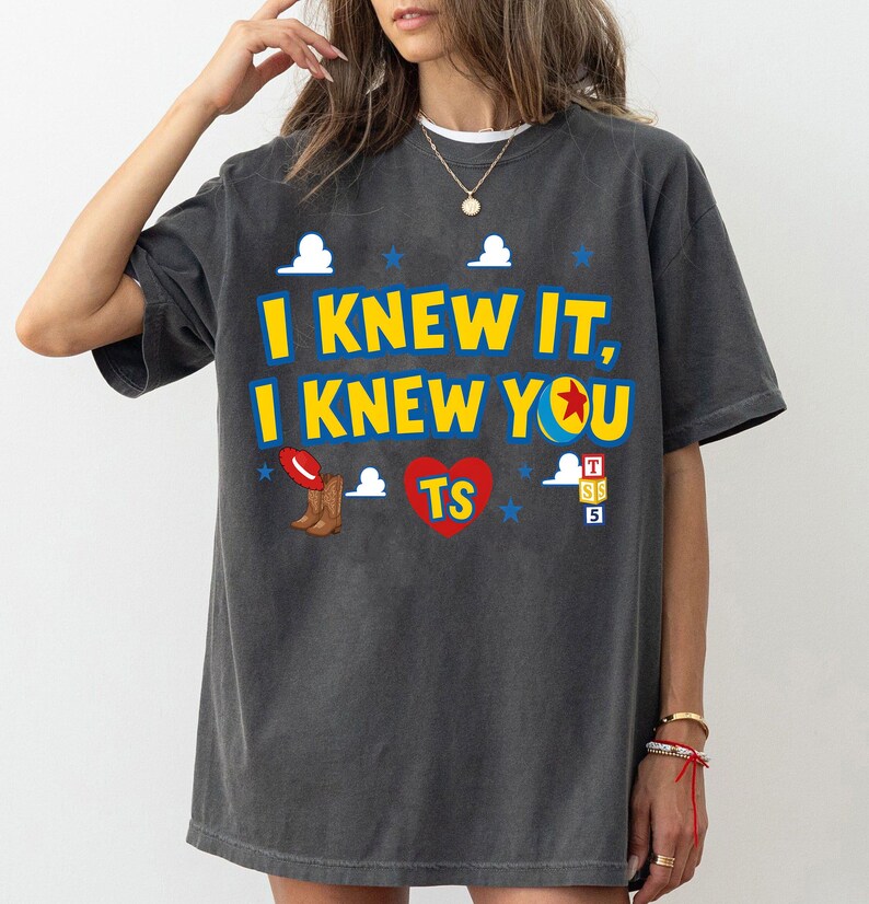 May include: A charcoal gray short-sleeve t-shirt featuring the yellow text I KNEW IT, I KNEW YOU with a ball icon, a red cowboy hat, brown boots, a red heart with TS, blue stars, white clouds, and a stack of alphabet blocks labeled T, S, 5.