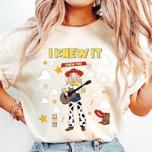 May include: A cream-colored t-shirt featuring a graphic of a cowgirl playing an acoustic guitar. The design includes the text "I KNEW IT", white clouds, yellow stars, musical notes, a red hat, brown boots, and a stack of blocks labeled "TSS5".
