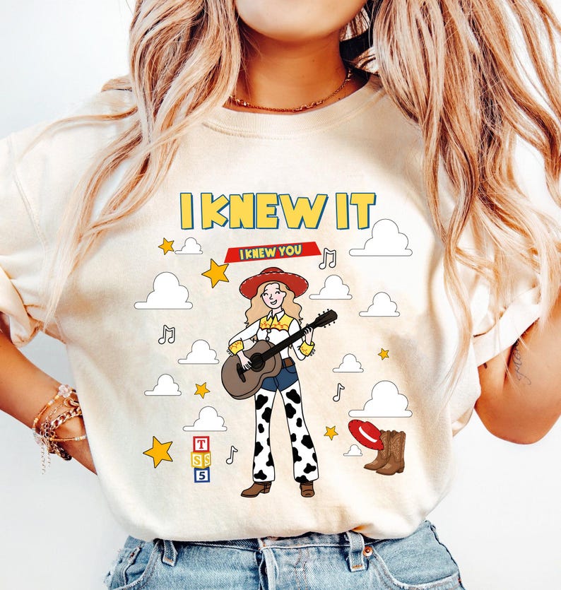 May include: A cream-colored t-shirt featuring a graphic of a cowgirl playing an acoustic guitar. The design includes the text "I KNEW IT", white clouds, yellow stars, musical notes, a red hat, brown boots, and a stack of blocks labeled "TSS5".